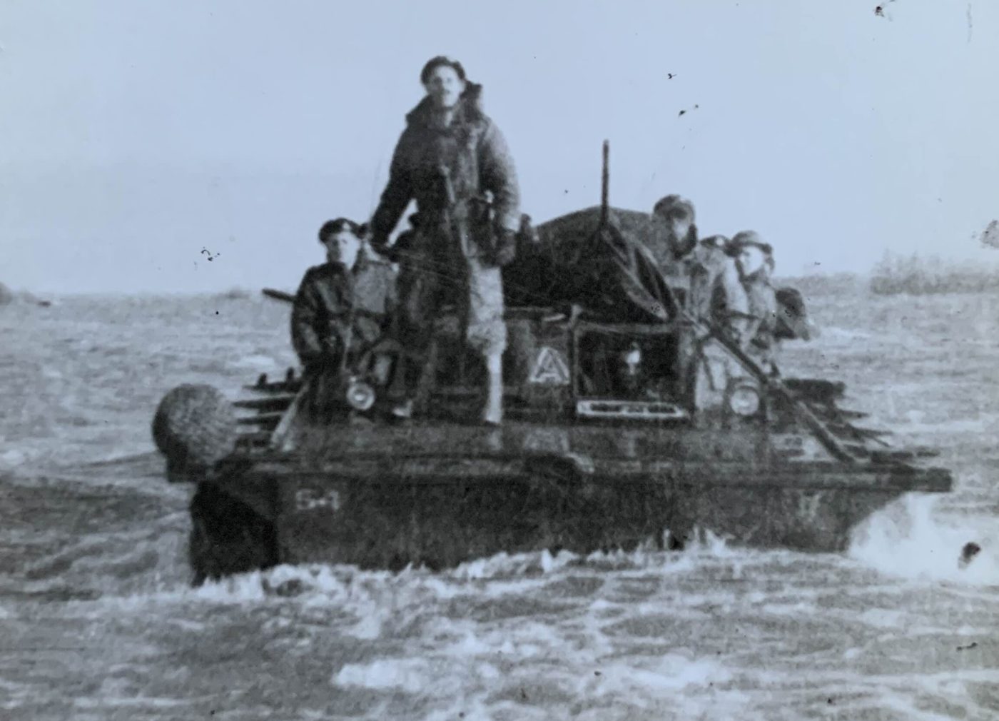 Soldiers aboard an amphibious vehicle crossing floodwater, one standing at the centre, in a black-and-white photograph from October 1944